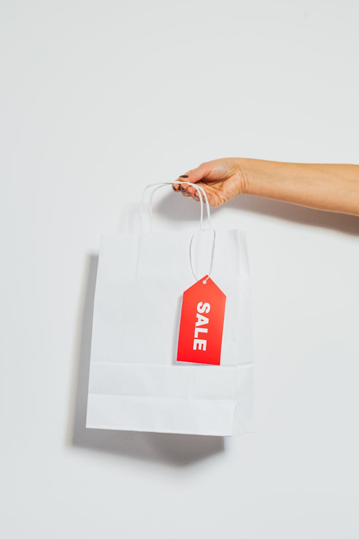 A person's hand holding a white shopping bag with a red sale tag on a white background.